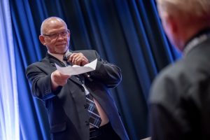 Photographers Of Las Vegas - Event Photography - Man in suit microphone pointing at crowd blue drapery